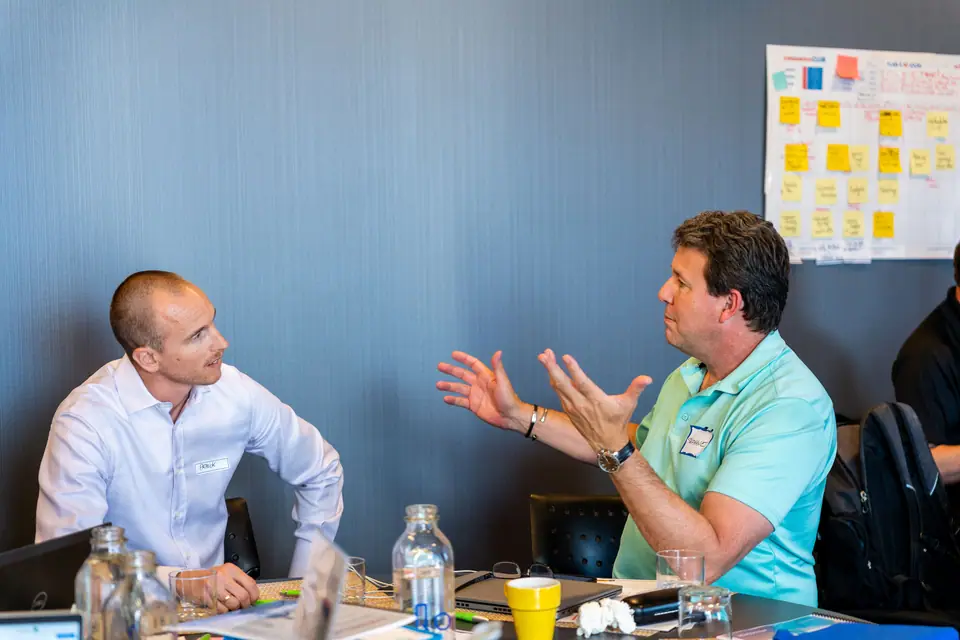 Deux entrepreneurs échangent autour d'une table, un plan d'action couvert de notes autocollantes au mur
